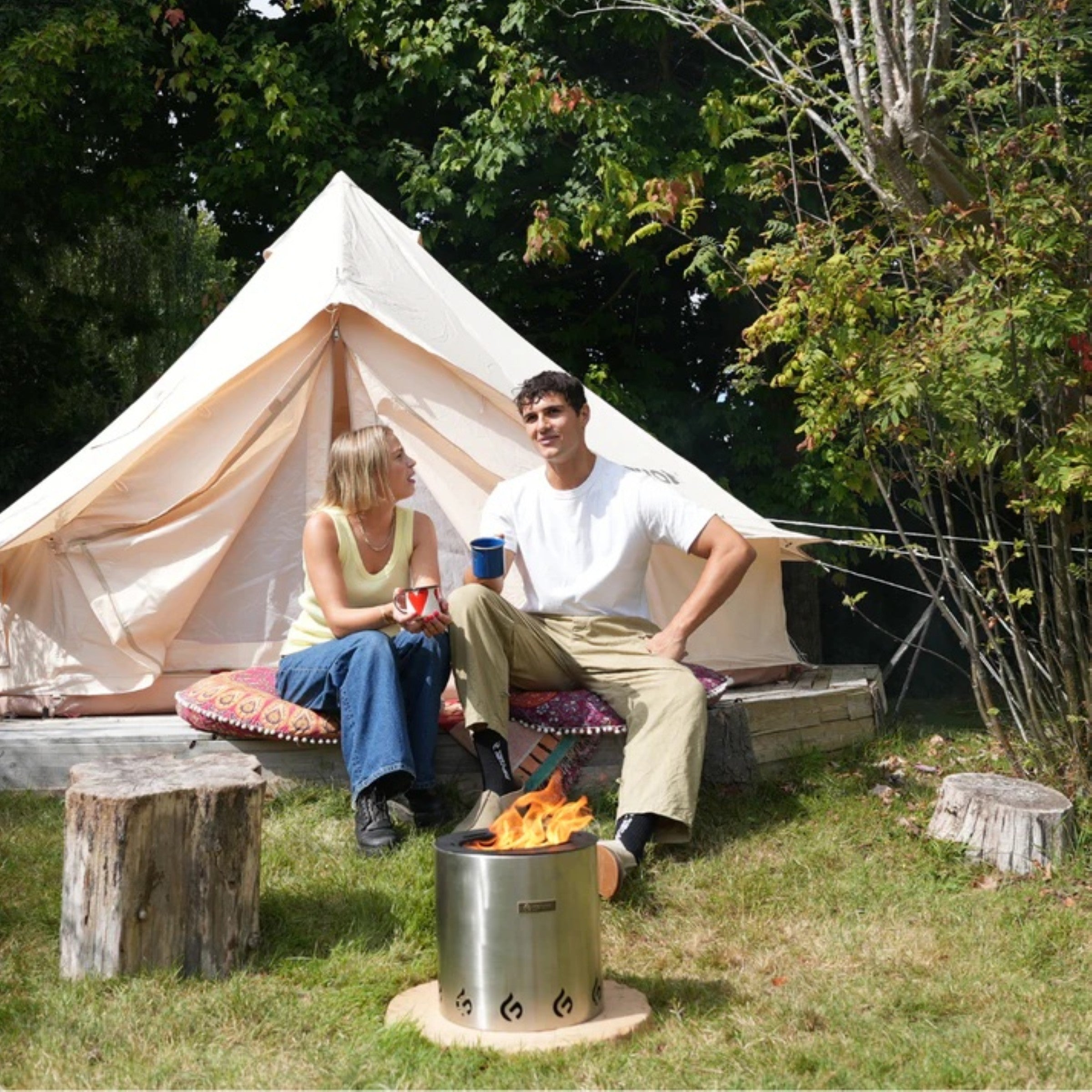 Two people sitting by a tent with a fire pit in a natural setting